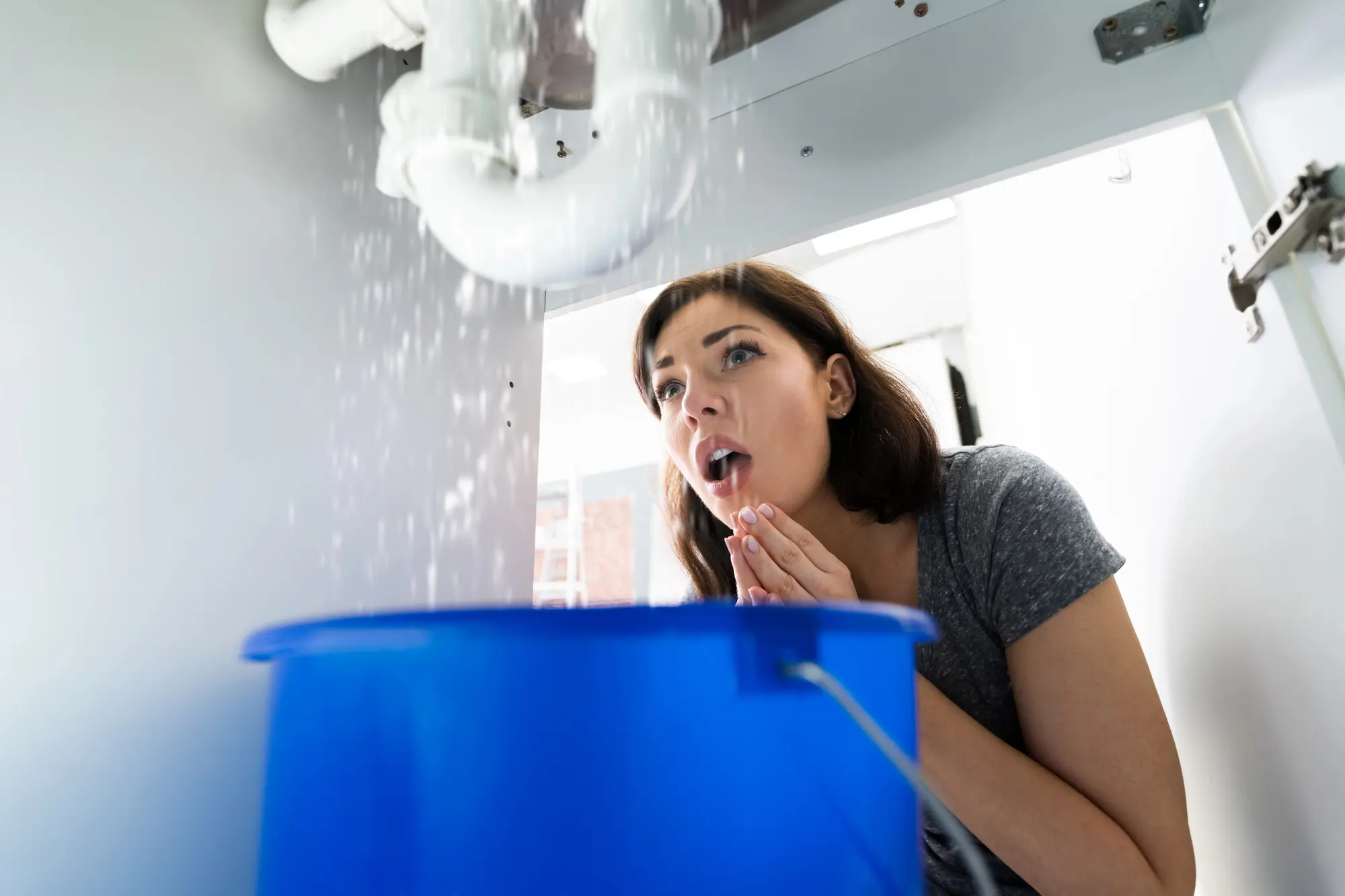 Homeowner looking up at ceiling in shock as water drips down, with blue bucket positioned to catch the leak